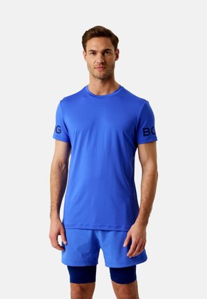 Homme portant un t-shirt de sport à manches courtes bleu vif avec du texte noir sur les manches, un short bleu assorti et des leggings de compression bleu foncé.