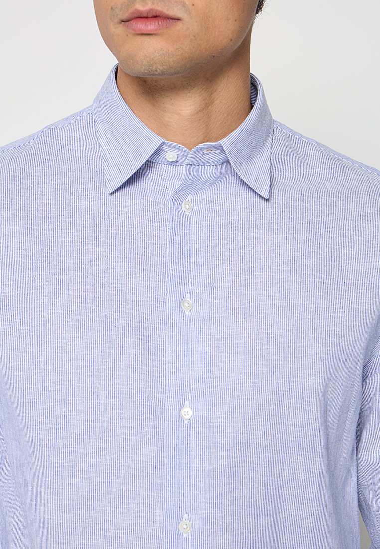 Hombre con camisa de rayas finas en azul claro y blanco, abotonada y con cuello abierto, visto desde el pecho hasta la parte inferior del rostro.