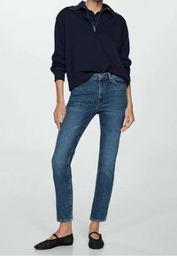 Marineblauer Pullover mit Viertelreißverschluss und gerippten Bündchen, kombiniert mit enganliegenden blauen Jeans und schwarzen Ballerinas. Glatte Stoffstruktur und schlankes Design.