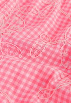 Rozā ginghemas audums ar izšūtiem ziedu rakstiem. Tekstūra ir gluda ar redzamām šuvēm un frēzētām detaļām ap zīmējumiem.