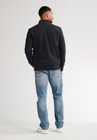 Felpa nera con colletto alto e finitura testurizzata, abbinata a jeans blu chiaro e sneaker bianche. Design minimalista, vestibilità moderna.