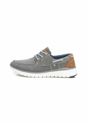 Zapatilla de tela gris con suela de goma blanca. Presenta acentos azules, detalles de cuero marrón y diseño con cordones. Textura suave y forma redondeada.