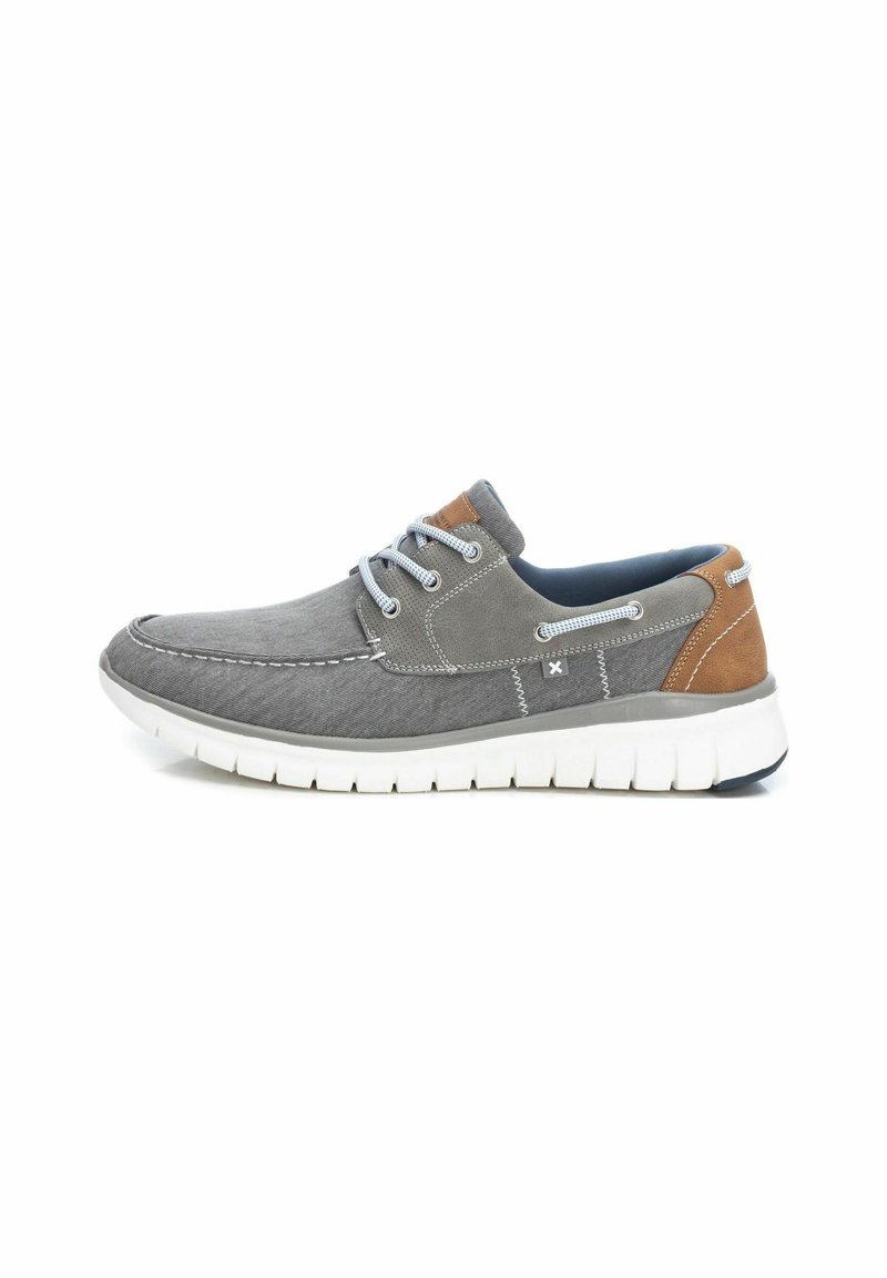 Zapatilla de tela gris con suela de goma blanca. Presenta acentos azules, detalles de cuero marrón y diseño con cordones. Textura suave y forma redondeada.