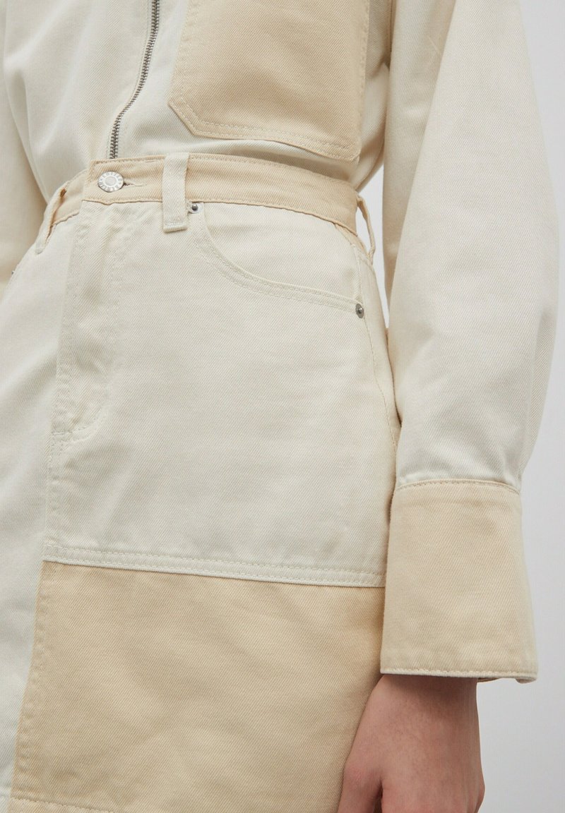 Beige en crème jumpsuit van katoen, met een ritsluiting, paneelontwerp en een knoopdetail in de taille. Lange mouwen met manchetten.
