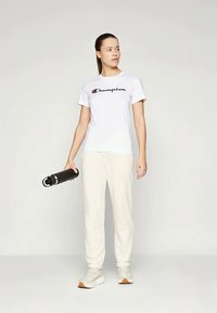 Witte katoenen t-shirt met "Champion" logo, lichtbeige joggingbroek en lichtgekleurde sneakers. Houdt een zwarte waterfles vast. Eenvoudige sportoutfit.