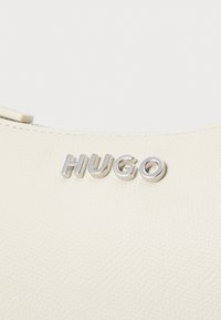 Vaalean beige-tekstuurinen käsilaukku, jossa on hopea "HUGO" -logo edessä, tyylikkäällä kaarevalla muotoilulla ja minimalistisella yksityiskohdalla.