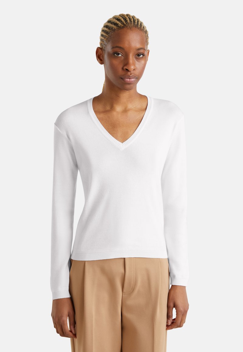 Maglione bianco a V in un tessuto morbido e liscio, con maniche lunghe e polsini a costine. Indossato su pantaloni beige, che mostrano una vestibilità casual.