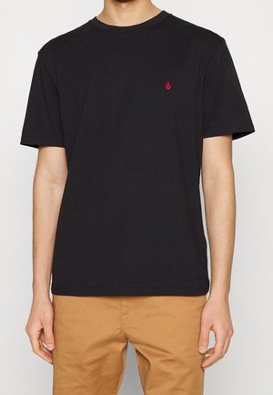 Person trägt ein schlichtes schwarzes T-Shirt mit kurzen Ärmeln und kleinem rotem Logo auf der Brust und hellbraune Hose, steht vor weißem Hintergrund.