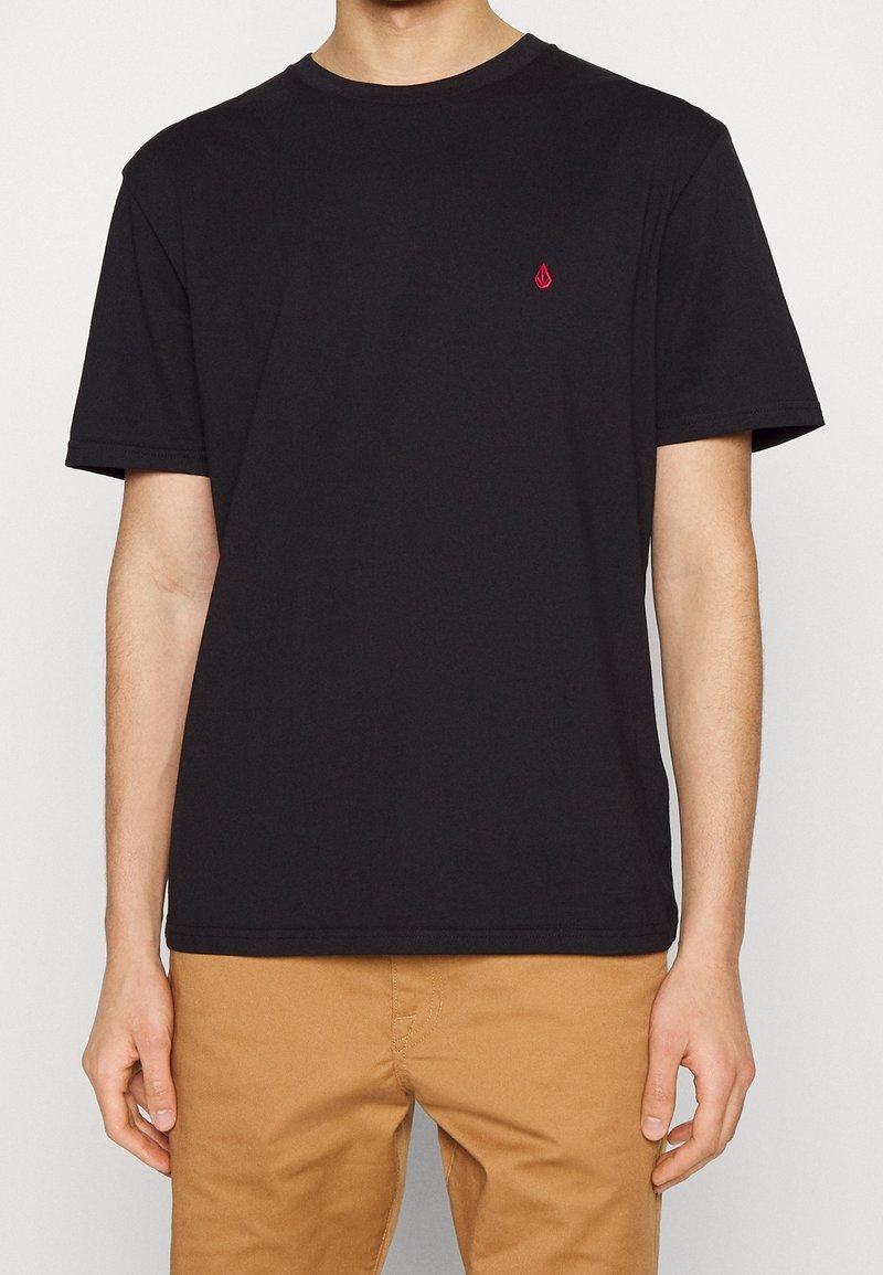 Personne portant un t-shirt noir uni à manches courtes avec un petit logo rouge sur la poitrine et un pantalon beige, debout devant un fond blanc.