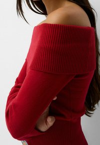 Femme aux longs cheveux noirs portant un pull côtelé rouge à épaules dénudées, les bras croisés et une main visible près de la taille.