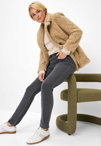Cappotto beige in tessuto peloso sopra un maglione chiaro, abbinato a jeans grigio scuro e sneakers bianche. Seduto su una sedia verde, curvilinea.