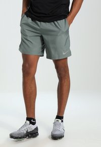 Šedé sportovní šortky s hladkou texturou a elastickým pasem, s malým logem Nike na levé noze. V kombinaci se šedými a černými teniskami.