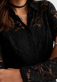 Svart spetstopp med blommönster, knappstängning och långa ärmar. Genomskinlig textur med en figursydd silhuett. Accenterad med ett choker-halsband.