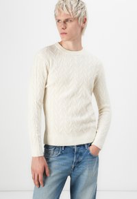 Cream-colored knitted sweater with a cable pattern, round neck, ribbed cuffs, worn with light blue denim jeans. Smooth texture.