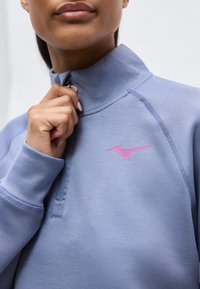 Gros plan sur une personne tirant la fermeture éclair d'un sweat-shirt col montant bleu clair avec un logo rose sur la poitrine.