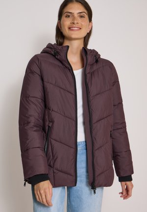 Femme portant une veste matelassée à capuche violet foncé par-dessus une chemise blanche et un jean bleu clair, souriant avec les mains détendues le long du corps.