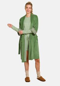 Grüner, weicher Stoffbademantel mit weiten Ärmeln, mit gemusterten Bündchen und zwei vorderen Taschen. Wird über einem passenden grünen Outfit getragen. Braune Hausschuhe.