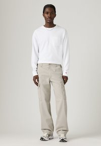 Biela košeľa s dlhým rukávom a vreckom na hrudi, skombinovaná s svetlo béžovými cargo nohavicami s bočnými vreckami a vzorovanými športovými topánkami.