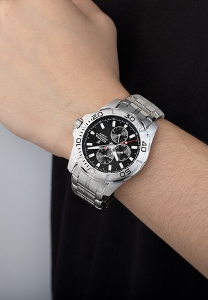 Montre en acier inoxydable argenté avec un cadran noir, trois sous-cadrans et de grands index numériques. Elle est dotée d'un bracelet en métal poli et d'un boîtier rond.