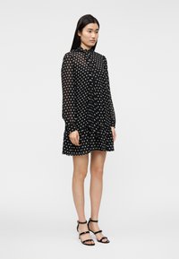 Γυναίκα που στέκεται φορώντας μαύρο διαφανές φόρεμα με μακριά μανίκια και λευκά πουά, καθώς και μαύρες ψηλοτάκουνες σανδάλες με λουράκια σε λευκό φόντο.