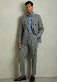 Sacou gri cu dungi fine, dublu rând, cămașă albastră, pantaloni gri cu model și mocasini negri, pe un fundal neutru.