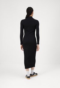 Vestito nero in maglia costina con maniche lunghe, silhouette aderente, lunghezza al polpaccio e collo alto. Indossato con calzini bianchi e scarpe da ginnastica nere.