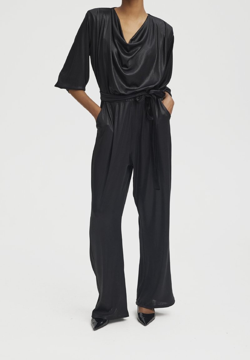 Femme portant une combinaison en satin noir avec un décolleté drapé, des manches trois-quarts, une ceinture à la taille, des poches et un pantalon à jambes larges avec des talons noirs pointus.