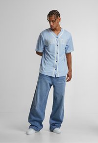 Camicia azzurra a maniche corte con bottoni, con bordi bianchi e gialli a contrasto, indossata con jeans in denim blu larghi e sneakers bianche.