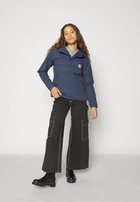 Marineblauer wasserdichter Mantel mit hohem Kragen, zwei vorderen Taschen und einem Logo-Patch, kombiniert mit weiten schwarzen Cargo-Hosen und schwarzen Stiefeletten.