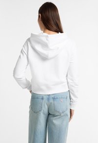 Felpa bianca cropped con cappuccio e polsini a coste, abbinata a jeans dritti azzurri chiari con tasche posteriori e un piccolo dettaglio del logo.