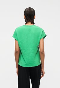 Άτομο με πλεκτά μαλλιά γυρισμένο ανάποδα, φορώντας ένα υφασμάτινο λαμπερό πράσινο μπλουζάκι με κοντά μανίκια και μαύρο παντελόνι, μπροστά σε απλό φόντο.