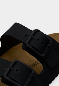 Κοντινό πλάνο από μαύρα λουριά σανδαλιών Birkenstock με ματ μαύρες αγκράφες πάνω σε καφέ πέλμα.