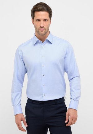 Man die een lichtblauw overhemd met knopen en donkere broek draagt, staand tegen een effen witte achtergrond.