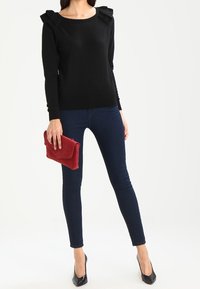 Zwarte trui met gerimpelde schouderaccenten, gecombineerd met donkerblauwe skinny jeans en zwarte hakken, terwijl ze een rode clutch vasthoudt.