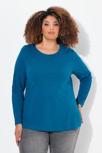 Donna con capelli ricci che indossa una maglietta a maniche lunghe color teal e pantaloni grigi, in piedi davanti a uno sfondo bianco con la mano sul fianco.