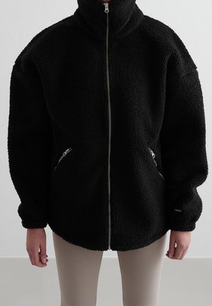 Osoba nosząca czarną, miękką kurtkę na zamek błyskawiczny z wysokim kołnierzem i zapinanymi na zamek bocznymi kieszeniami, połączoną z jasnobeżowymi spodniami.