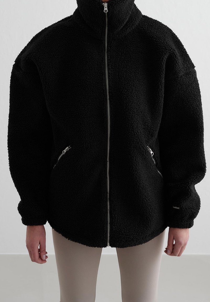 Personne portant une veste noire duveteuse à fermeture éclair avec col montant et poches latérales zippées, associée à un pantalon beige clair.