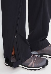 Bas des jambes portant un pantalon noir avec des fermetures éclair latérales ouvertes au-dessus de chaussures de trail grises et roses.