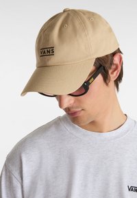 Gorra de béisbol de color beige claro con un logo negro "VANS" en la parte frontal, con una corona redondeada y visera curva, llevada con una camiseta gris.