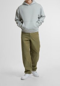 Person som bär en ljusgrå hoodie, olivgröna byxor och vita sneakers, stående med händerna i hoodiefickan.