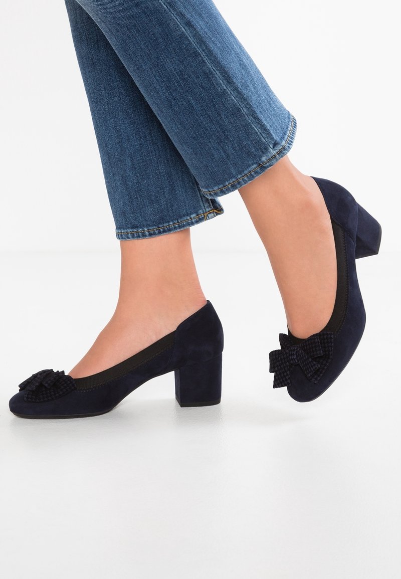 Pieds chaussés de chaussures à talons blocs en daim bleu foncé avec des nœuds texturés, associés à un jean denim bleu, sur un fond blanc.