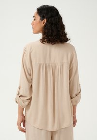 Beige Bluse mit langen Ärmeln, entspannter Passform und dezentem Rückenfalten. Weicher Stoff, aufgeschlagene Manschetten und abgerundeter Saum.