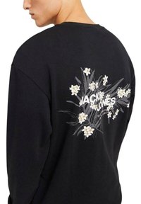 Sudadera negra con un estampado gráfico floral en blanco y amarillo, con el texto "JACK & JONES" y un corte relajado. Textura de tela suave.