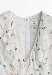 Robe florale blanche avec encolure froncée. Présente de petites fleurs roses et bleues, une fermeture éclair dans le dos, et un tissu léger et texturé.