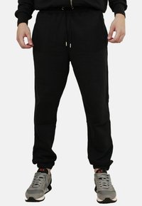 Pantaloni da jogging neri con vita elasticizzata, chiusura con cordino, tasche laterali e polsini aderenti, abbinati a scarpe da ginnastica grigie.