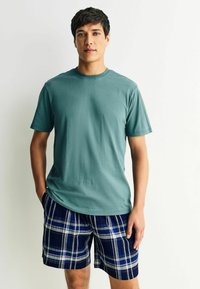 Lyhythihainen teal-vihreä puuvillapaita, joka on yhdistetty laivastonsinisiin ruudullisiin shortseihin, joissa on sinisiä ja valkoisia raitoja. Paidassa on pyöreä pääntie ja rento istuvuus.