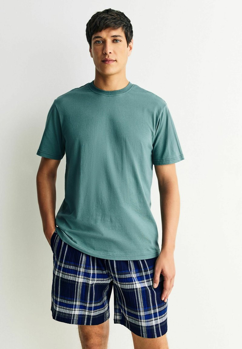 Lyhythihainen teal-vihreä puuvillapaita, joka on yhdistetty laivastonsinisiin ruudullisiin shortseihin, joissa on sinisiä ja valkoisia raitoja. Paidassa on pyöreä pääntie ja rento istuvuus.