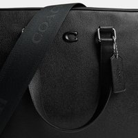 Musta teksturoitu nahkalaukku, jossa leveä olkahihna, metallinen solki ja nahkapala, joka roikkuu ketjussa kahvassa.