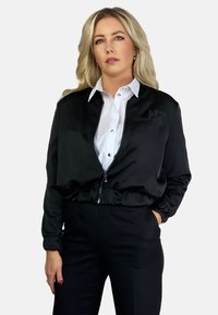 Czarna kurtka bombowca z gładkiego satynowego materiału, z zamkiem z przodu, elastycznym pasem i ściągaczami przy rękawach, w zestawie z białą koszulą z kołnierzykiem.