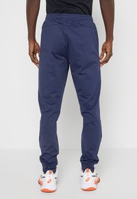 Granatowe spodnie sportowe z elastycznym paskiem w talii i zwężającymi się nogawkami, charakteryzujące się gładką fakturą i brakiem widocznych wzorów. Noszone z białymi butami sportowymi.